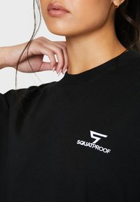 Sort t-shirt lavet af bomuld, med korte ærmer, rund halsudskæring og hvid "SQUATPROOF" logo på brystet, glat tekstur.
