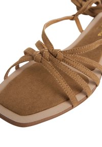 Brun mockasandal med flera tunna remmar, en platt design och fyrkantig tåform. Innersulan är lätt vadderad för komfort.