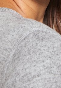 Vaaleanharmaa neulepusero, jossa on teksturoitu kuvio ja resorireunukset kaula-aukossa ja hihansuissa, korostaen pehmeää kangasta ja rentoa suunnittelua.