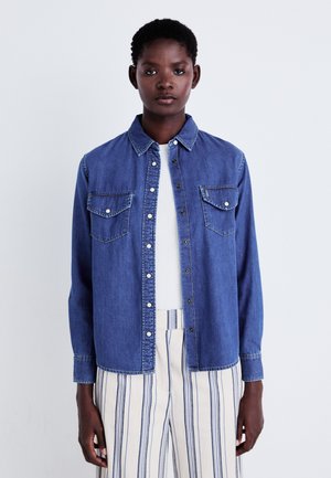Jeanshemd in tiefblauer Farbe, mit Knopfleiste und zwei Brusttaschen, klassischer Kragen, lange Ärmel, kombiniert mit gestreiften Hosen.