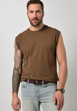 Mann mit gestutztem Bart, der ein braunes ärmelloses Hemd, hellblaue Jeans, Ledergürtel, Uhr und ein detailliertes Tätowierarmband am linken Arm trägt.