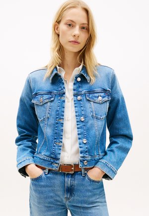 Niebieska kurtka dżinsowa o klasycznym kroju, z przednimi kieszeniami, srebrnymi guzikami i przeszyciami, zestawiona z białą koszulą i brązowym paskiem.