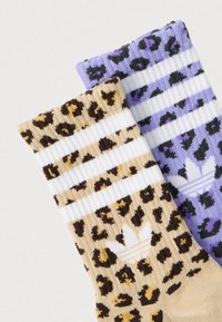 Divi pāri zeķu ar leopardzīmējumu, viens bēšā krāsā un otrs purpursarkanā, katrām ar baltām svītrām un baltu Adidas logotipu pie potītes.