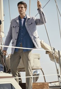 Hellgraue Jacke mit mehreren Taschen über einem blauen Hemd mit Knöpfen. Neutrale Cargohosen, stehend auf einem Boot mit Segelausrüstung im Hintergrund.