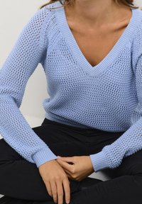 Persona che indossa un maglione azzurro chiaro a V in maglia e pantaloni neri, seduta con le mani appoggiate sulle gambe incrociate su uno sfondo uniforme.