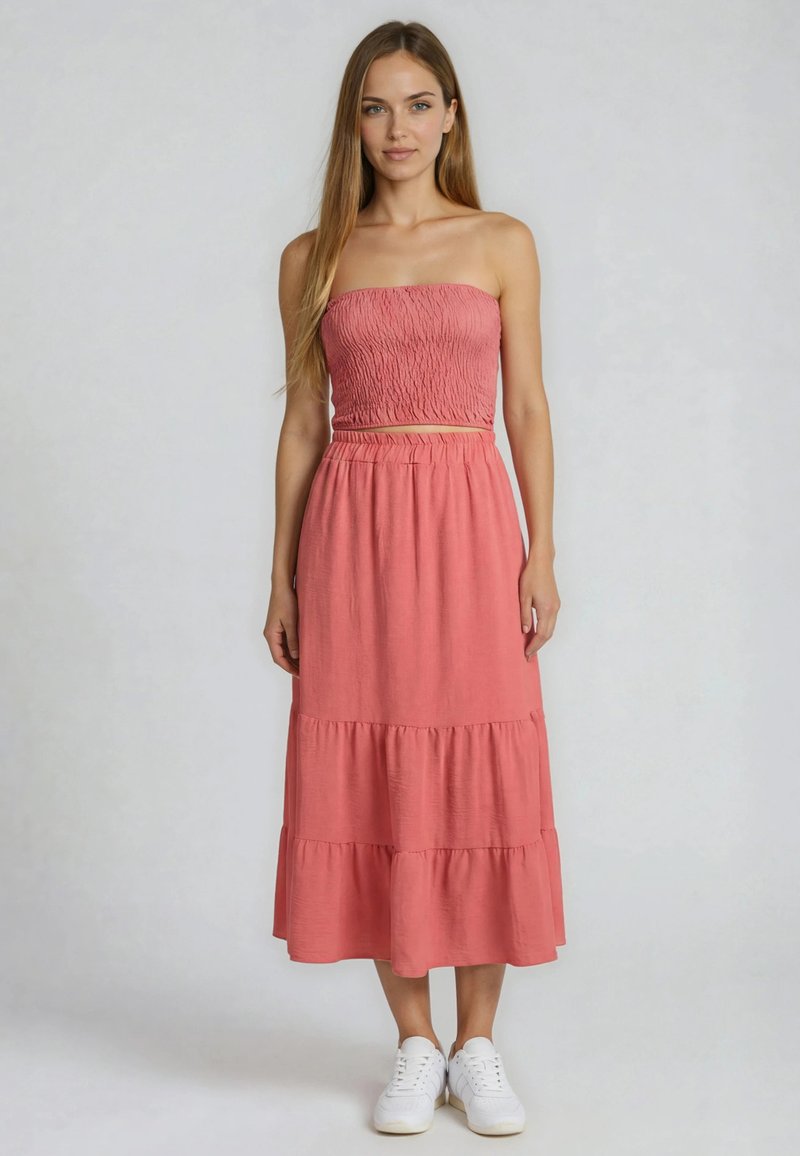 Jeune femme portant un crop top rose smocké sans manches et une jupe à volants assortie, debout avec des baskets blanches sur un fond uni.