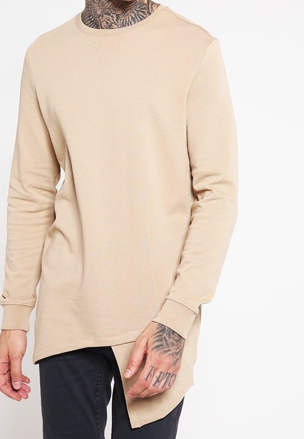 Chemise beige à manches longues avec un ourlet asymétrique portée par une personne tatouée, associée à un pantalon noir.