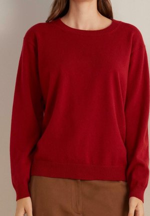 Kvinde iført en ensfarvet rød langærmet sweater stoppet ned i brune bukser, står mod en neutral baggrund.