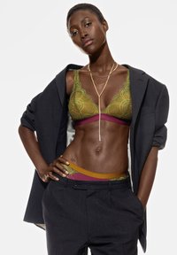 Grön spets-bh med intrikata mönster, kombinerad med ett vinrött band och gula detaljer synliga ovanför skräddarsydda grå byxor. Guldkedja.