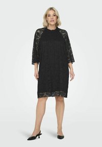 Czarna koronkowa sukienka z rękawami o długości trzech ćwierć, charakteryzująca się luźnym krojem w kształcie litery A oraz misternymi wzorami kwiatowymi. Noszona z czarnymi szpilkami.
