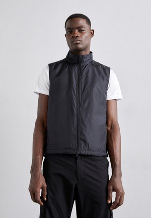 Gilet nero senza maniche in un tessuto liscio e leggero, con colletto alto e zip frontale, abbinato a una t-shirt bianca sottostante.