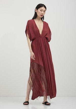 Femme portant une robe maxi rouge profond avec un décolleté plongeant et des franges sur un côté, associée à des sandales noires à enfiler.