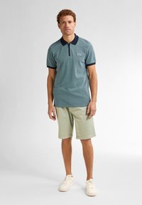 Cămașă polo texturată de culoare albastru deschis cu guler bleumarin și închidere cu fermoar, asortată cu pantaloni scurți bej și adidași crem.