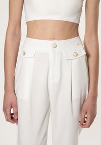 Witte linnen broek met een hoge taille, twee gouden knopen en ingesneden zakken. De stof lijkt lichtgewicht met een subtiele textuur.