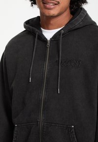 Sudadera negra con cremallera y tejido texturizado, que presenta un bolsillo canguro, capucha con cordón y detalle bordado en el pecho.