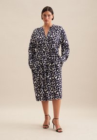 Abito blu navy e bianco con scollo a V, vita con cintura, maniche lunghe e tasche laterali, caratterizzato da un motivo a pois e una lunghezza fino al ginocchio.