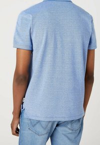 Polo à manches courtes en tissu bleu clair chiné avec un col, des fentes sur les côtés et une étiquette sur l'ourlet. Assorti à un jean en denim bleu clair.
