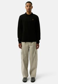 Czarna koszulka polo z długim rękawem i złotym logo, połączona z beżowymi spodniami cargo z bocznymi kieszeniami i szarymi trampkami.