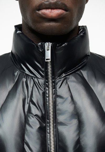 Κοντινό πλάνο ενός ατόμου που φοράει μια γυαλιστερή μαύρη κρύα μπουφάν με ασημένιο κεντρικό φερμουάρ κλειστό μέχρι τον λαιμό.