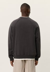 Άνδρας που στέκεται με την πλάτη στην κάμερα, φορώντας σκούρο γκρι φούτερ και ανοιχτό μπεζ παντελόνι, με απλό φόντο.