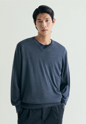 Ung mand iført en mørkegrå V-hals sweater over en navy skjorte, stående med hænderne i lommerne mod en ensartet lys baggrund.