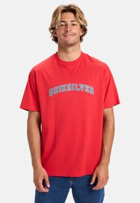 Camiseta de algodón roja con un logo curvado en azul "QUIKSILVER" en el pecho, mangas cortas y un ajuste relajado.