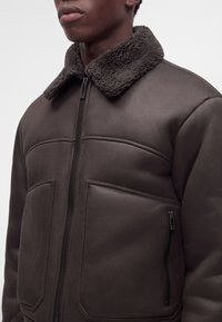 Homme portant une veste marron foncé avec un col en peau lainée, une fermeture éclair devant et de grandes poches avant, visible des épaules au milieu du torse.