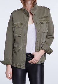 Veste militaire de couleur vert olive avec col montant, boutons pression et deux poches poitrine, portée sur un haut blanc et un pantalon en cuir noir.