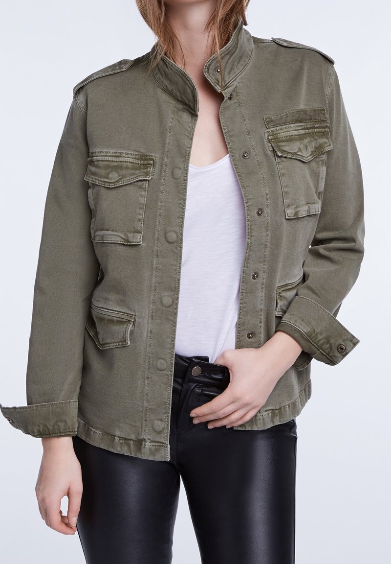 Veste militaire de couleur vert olive avec col montant, boutons pression et deux poches poitrine, portée sur un haut blanc et un pantalon en cuir noir.