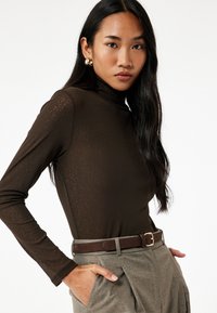 Ruskea pitkähiha turtleneck-paita, jossa on tekstuurinen pinta, yhdistettynä harmaisiin korkea vyötäröisiin housuihin ja ruskeaan vyöhön, jossa on kultainen solki.