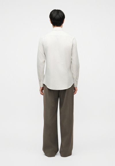 Person som har på seg en langermet hvit skjorte og løstsittende brune bukser, stående med ryggen mot kameraet foran en ensfarget hvit bakgrunn.