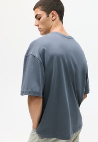 Šedé tričko s krátkým rukávem, uvolněného střihu a kulatým výstřihem. Měkká tkanina s hladkým povrchem, zobrazené zezadu, ruce v kapsách.