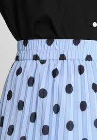 Una gonna plissettata blu chiaro con pois neri, dotata di una vita elastica e di una texture liscia. Parte di un top nero è parzialmente visibile sopra.