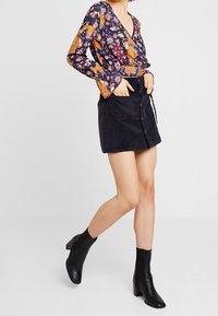 Bloemrijke longsleeve wikkeltop in marineblauw, oranje en geel; gecombineerd met een zwarte corduroy minirok met frontknopen en zijzakken.