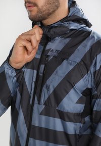 Veste à capuche résistante à l'eau en noir et bleu avec motif géométrique, dotée d'une poche zippée et de poignets élastiques pour un ajustement modulable.