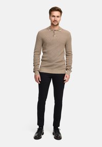 Polo beige a maniche lunghe con un tessuto lavorato, apertura a tre bottoni, abbinato a pantaloni slim-fit neri e scarpe eleganti nere.