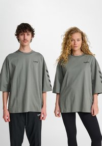 Twee personen dragen oversized, groene katoenen t-shirts met zwarte logo- en streepdetails. De een combineert het met zwarte broek, de ander met leggings.