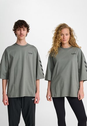 Twee personen dragen oversized, groene katoenen t-shirts met zwarte logo- en streepdetails. De een combineert het met zwarte broek, de ander met leggings.