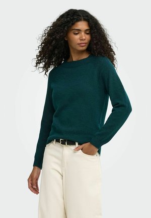 Femme aux cheveux bouclés portant un pull vert foncé à manches longues et un pantalon beige clair, debout avec une main dans sa poche.