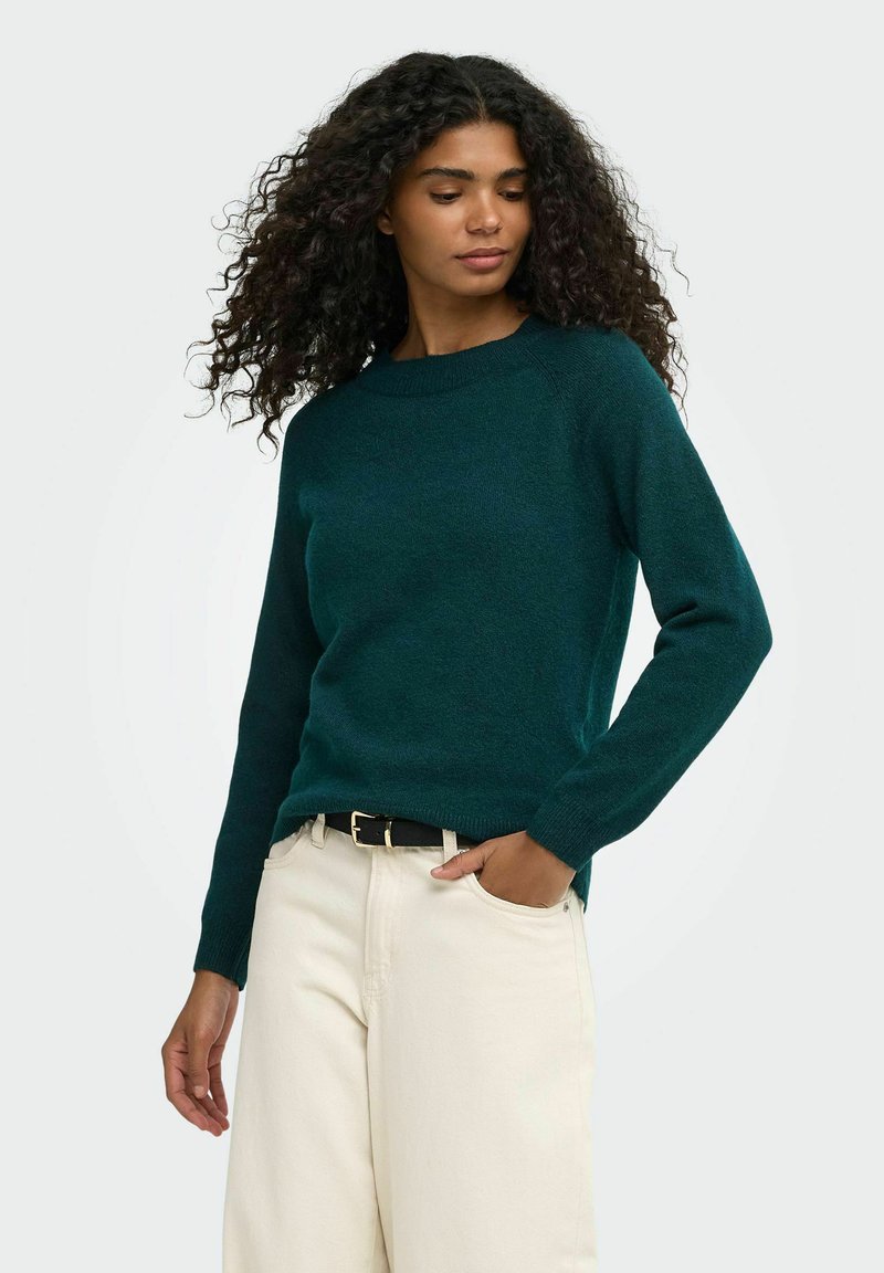 Femme aux cheveux bouclés portant un pull vert foncé à manches longues et un pantalon beige clair, debout avec une main dans sa poche.