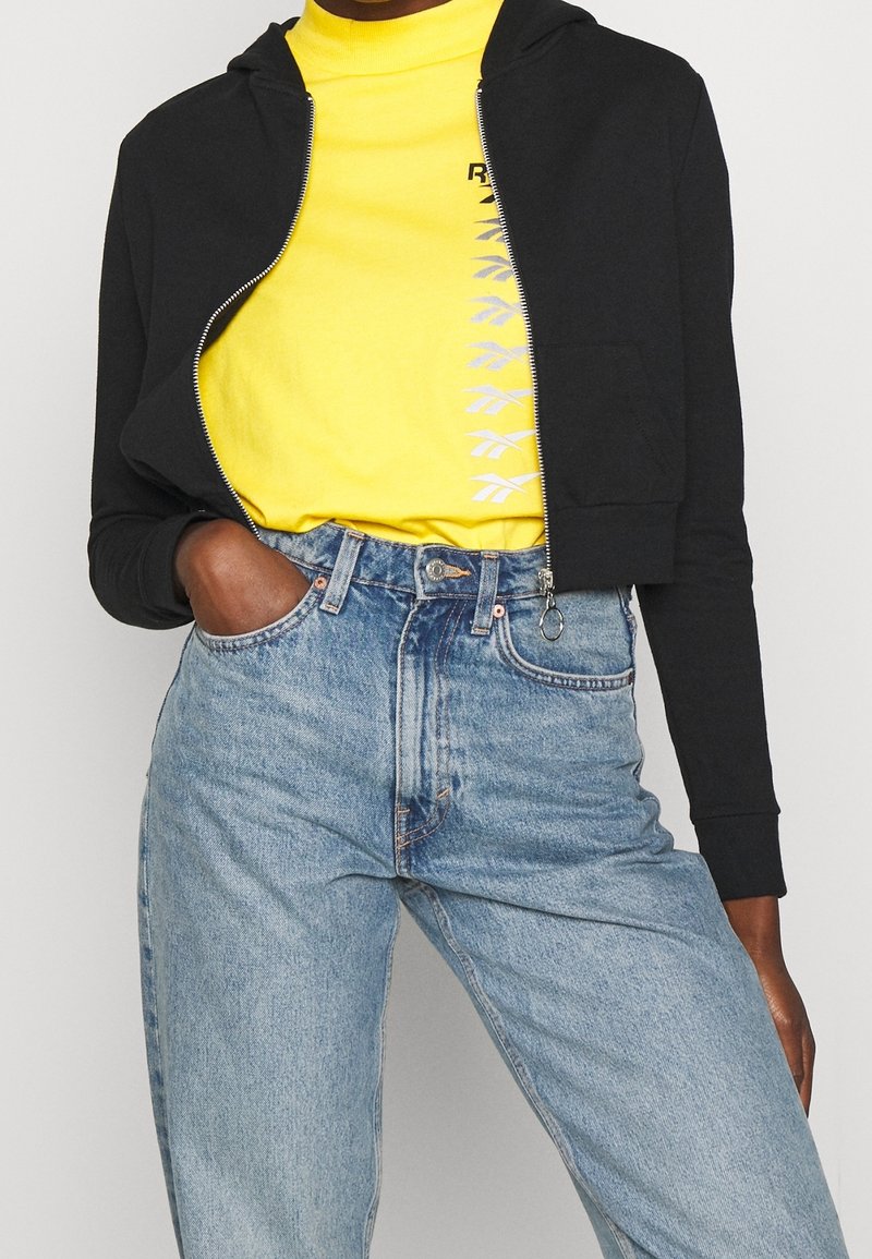 Personne portant un sweat à capuche court noir zippé, un t-shirt jaune vif avec un logo vertical, et un jean bleu clair taille haute, main dans la poche.
