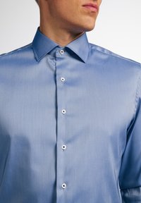 Uomo che indossa una camicia elegante azzurro chiaro con colletto alla francese, mostrato dal collo fino a metà torso, rivolto leggermente verso destra.