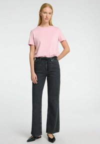 Camiseta de algodón rosa claro, jeans de mezclilla oscuros de corte recto, zapatos negros de slip-on, de pie contra un fondo neutro.