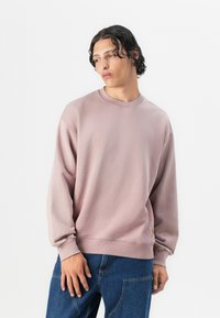 Lys rosa genser med lange ermer og ribbet rundhals. Har et lite logomerke på den nedre siden. Myk tekstur, avslappet passform.