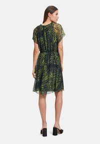 Grünes und schwarzes transparentes Kleid mit Zebra-Muster, weiten Ärmeln, tailliertem Bund und einem fließenden Rock. Kombiniert mit schwarzen klobigen Absätzen.
