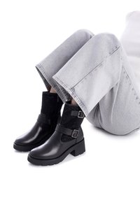 Botas de cuero negras hasta el tobillo con una sección de tela negra texturizada, con cremallera lateral, correas y un tacón cuadrado. Combinadas con pantalones grises cortos.