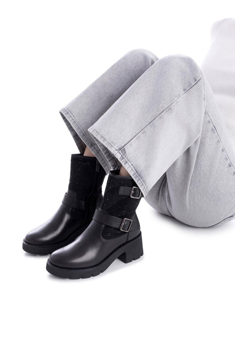 Botas de cuero negras hasta el tobillo con una sección de tela negra texturizada, con cremallera lateral, correas y un tacón cuadrado. Combinadas con pantalones grises cortos.