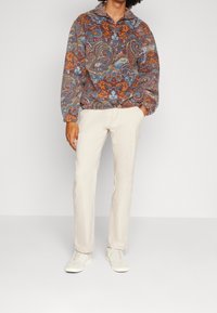 Pullover in pile con motivo paisley in marrone, blu e arancione. Accoppiato con pantaloni beige e sneakers grigie, con colletto zip.