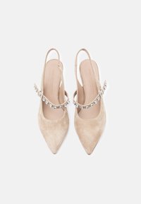 Μπεζ καστόρινα τακούνια με λουράκι στο πίσω μέρος και μυτερή μύτη. Διαθέτουν διακοσμητικό λουράκι με ασημί διακοσμητικά. Λείο εσωτερικό επένδυση και μινιμαλιστικός σχεδιασμός.