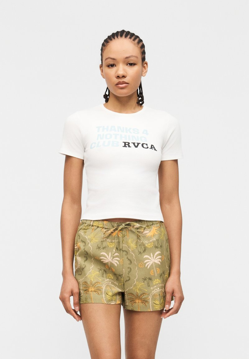 Νεαρή γυναίκα με πλεξούδες φοράει ένα λευκό crop top με κείμενο και πράσινα σορτς με σχέδια από φοίνικες και τροπικά μοτίβα.